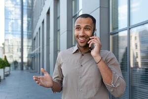 alegre e sorridente africano americano escritório trabalhador falando em a telefone, homem lado de fora escritório construção dentro camisa caminhando dentro a cidade, homem de negocios em almoço pausa foto