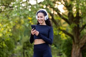 jovem lindo latim americano mulher anda em dentro a parque durante ativo fisica exercícios, mulher usa a inscrição para ouvindo para audio livros e música em telefone, sorridente desportista dentro fones de ouvido. foto