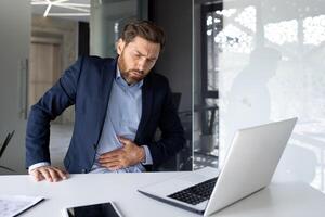 uma jovem masculino homem de negocios é sentado dentro a escritório às uma escrivaninha com uma computador portátil e detém dele estômago com dele mãos torcido. sente dor e desconforto. foto