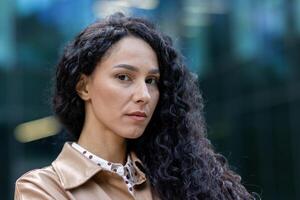 retrato do maduro adulto o negócio mulher lado de fora escritório prédio, fechar acima latim americano mulher com encaracolado cabelo olhando às Câmera, mulher patrão financista dentro o negócio roupas. foto