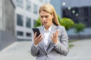 fechar-se do chateado Senior o negócio mulher segurando telefone, fêmea trabalhador dentro o negócio terno recebendo mau acusações on-line, patrão usando aplicativo em Smartphone, caminhando lado de fora escritório construção dentro cidade. foto