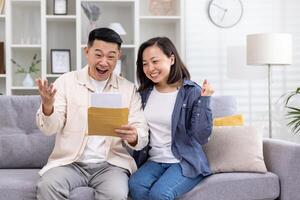 alegre e feliz ásia família junto, homem e mulher às casa em sofá sentado dentro vivo sala, recebido postal envelope com Boa notícias, mensagem e Boa realização notícias. foto