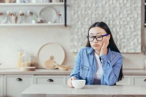café pausa. jovem cansado ásia mulher trabalhador autonomo. sentado às casa às a cozinha mesa com uma copo do café, em repouso, considerado. foto