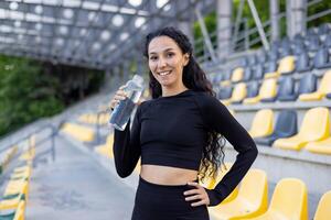 uma em forma hispânico mulher pausando para beber água durante dela exercício rotina dentro a esvaziar estádio. foto