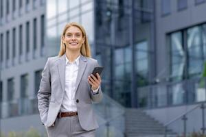 retrato do uma jovem empresária dentro uma terno caminhando lado de fora a escritório Centro, segurando uma telefone e mão dentro dela bolso, sorridente e olhando às a Câmera. foto