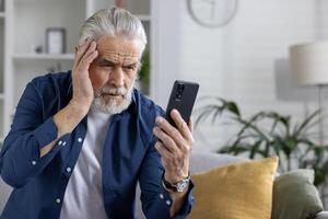 preocupado Senior homem às casa olhando às dele telefone, exibindo emoções do recebendo infeliz ou mau notícias, segurando cabeça dentro sofrimento. foto