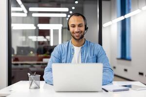 uma alegre masculino cliente serviço representante com fones de ouvido trabalhando em uma computador portátil dentro uma moderno escritório contexto. foto