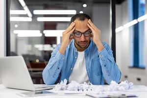jovem hispânico homem sentado chateado às a escrivaninha dentro a escritório, segurando dele cabeça dentro frustração, amassado documentos e papel. foto