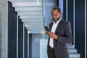 bem sucedido africano americano homem de negocios dentro escritório em pé de janela, patrão usando telefone, homem digitando mensagem sorridente e lendo conectados notícias, digitando mensagem dentro social rede aplicativo. foto