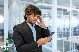 chateado hispânico homem dentro escritório às trabalhos lendo mau notícia a partir de telefone on-line, homem de negocios dentro camisa perto janela trabalhando dentro escritório, usando Smartphone. foto