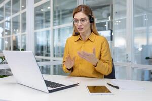 maduro adulto o negócio mulher com loiro cabelo trabalhando dentro luz escritório, cliente serviço tecnologia trabalhador sorridente e falando para cliente consultando, usando fone de ouvido e computador portátil para controlo remoto chamar. foto