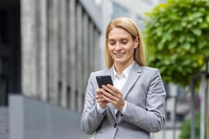 bem sucedido mulher patrão anda em lado de fora escritório construção ao ar livre, dentro o negócio terno, empresária usa aplicativo em telefone, sorrisos contente lê Internet Páginas, navega notícias, escreve texto mensagem, chamar. foto