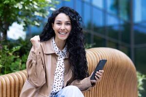 jovem atraente hispânico mulher com encaracolado cabelo sentado em Banco lado de fora segurando telefone dentro mão e sorridente olhando às Câmera, feliz morena dentro excitação do ganhando on-line, obtendo Boa notícias. foto