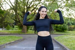 uma alegre hispânico mulher flexiona dela músculos com confiança dentro uma parque, exibindo força e ginástica dentro uma natural contexto. vestido dentro desportivo Preto traje, ela sorrisos amplamente. foto