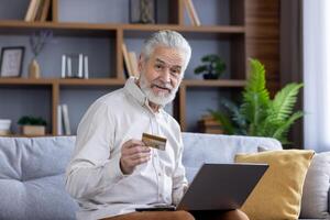 confiante Senior homem segurando uma crédito cartão enquanto usando uma computador portátil em uma sofá às lar, exibindo a facilidade do conectados compras. foto