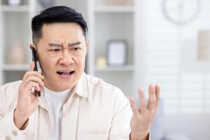 Bravo Bravo e chateado homem falando em telefone às lar, ásia trabalhando remotamente a partir de casa escritório sentado às mesa dentro vivo sala, freelance homem de negocios às trabalhar. foto