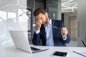 uma cansado jovem homem, uma homem de negocios, uma advogado senta dentro a escritório às uma mesa, esfrega dele olhos, detém copos, descansa. foto