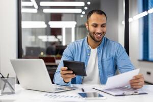 focado indiano homem de negocios dentro uma moderno escritório contexto, trabalhando em dele computador portátil enquanto consultando documentos em uma escrivaninha. foto