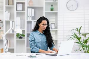 sério hispânico empresária trabalhando em dela computador portátil dentro uma ordenadamente organizado casa escritório com uma branco moderno decoração. foto