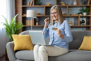 alegre mulher sentado em sofá com computador portátil, levantando punho enquanto segurando uma crédito cartão, sentindo-me vitorioso depois de a conectados transação. foto