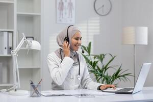 alegre fêmea médico dentro hijab sentado às dela escrivaninha dentro uma moderno clínica, fornecendo consulta através da fone de ouvido e computador portátil, com uma caloroso sorriso. foto