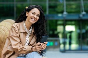 mulher noivando com dela Smartphone enquanto sentado em uma público Banco no meio uma moderno cidade pano de fundo. isto imagem transmite conectividade, tecnologia dentro todo dia vida, e a mistura do estilo de vida e digital noivado. foto