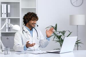 jovem indiano médico consultando pacientes remotamente, homem dentro branco médico casaco com fone de ouvido usando computador portátil para chamar, sorridente contente dentro clínica escritório. foto