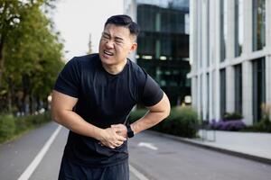 jovem ásia masculino desportista em pé em uma cidade rua e segurando dele estômago com dele mãos. sente e caretas a partir de forte dor depois de Treinamento e corrida. foto