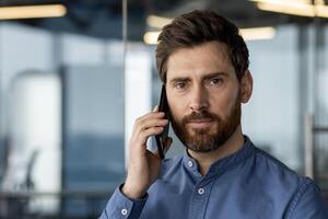 fechar-se retrato do uma jovem homem dentro uma azul camisa em pé dentro uma moderno escritório e falando em a telefone, olhando com confiança e a sério para dentro a Câmera. foto