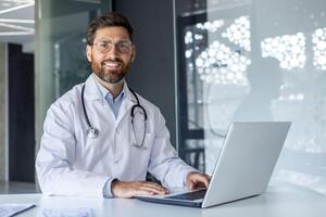 retrato do uma jovem sorridente masculino médico sentado dentro a escritório do a clínica às a trabalhando mesa dentro uma branco casaco, e trabalhando em uma computador portátil. foto