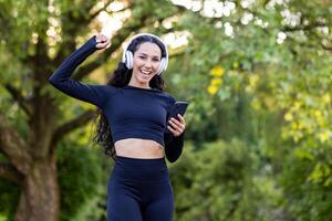 feliz mulher dentro Preto roupa ativa e branco fones de ouvido levantando mão com cerrado punho enquanto usando Smartphone. desportivo fêmea com encaracolado cabelo alcançando objetivo do corrida maratona em público parque fundo. foto