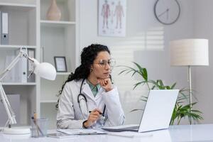 focado hispânico fêmea médico sentado às dela escritório mesa, trabalhando em uma computador portátil com papelada e médico equipamento em volta. foto