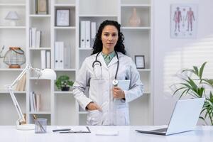retrato do sério confiante fêmea médico dentro médico escritório do hospital, hispânico mulher dentro branco médico casaco em pé perto mesa, segurando tábua computador, olhando às Câmera. foto