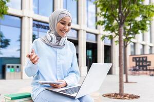 focado jovem aluna vestindo uma hijab com fones de ouvido estudando em dela computador portátil e levando notas lado de fora em campus. foto