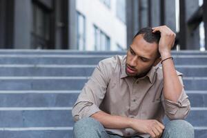 desapontado e triste homem sentado em escadas lado de fora escritório prédio, empregado disparamos a partir de trabalhar, africano americano homem dentro camisa chateado e depressivo foto