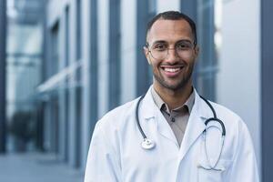 fechar-se foto retrato do jovem africano americano médico lado de fora hospital, homem dentro médico casaco sorridente e olhando às Câmera