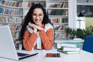 jovem hispânico mulher estudando dentro acadêmico universidade biblioteca, fêmea aluna sorridente e olhando às Câmera enquanto sentado às computador portátil, mulher com encaracolado cabelo. foto