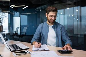 bem sucedido financista investidor trabalho com documentos e contas dentro escritório, homem de negocios com barba sorridente satisfeito com trabalhos resultados e conquistas, homem dentro escritório com computador portátil trabalhando. foto