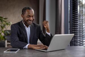 uma homem dentro uma terno é sentado às uma escrivaninha com uma computador portátil e uma tábua. ele é sorridente e ele é feliz foto