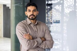 retrato do uma confiante homem dentro o negócio casual vestuário cruzando dele braços com uma dica do uma sorrir dentro a escritório. foto