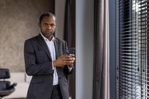 a africano americano homem de negocios Verificações dele telefone de a janela dentro uma moderno escritório configurar, exalando uma profissional e focado atmosfera. foto