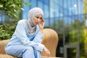 uma muçulmano mulher dentro uma hijab parece perturbado ou contemplativo enquanto sentado sozinho em uma parque Banco fora. foto