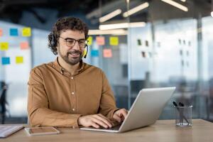 alegre masculino profissional usando computador portátil e vestindo uma fone de ouvido dentro uma moderno escritório ambiente, exibindo positividade e produtividade. foto