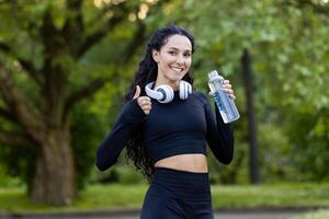 uma alegre hispânico mulher goza uma ginástica pausa às a parque, segurando uma água garrafa e vestindo fones de ouvido, dando uma polegares acima com árvores dentro a fundo. foto