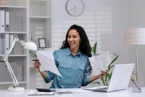 alegre hispânico mulher revendo papéis com uma sorrir dentro dela bem organizado casa escritório contexto, incorporando profissional conforto e produtividade. foto