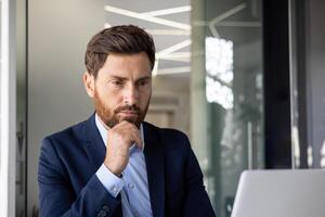 fechar-se foto do uma jovem sério masculino homem de negocios sentado focado dentro a escritório às uma computador portátil e pensando sobre Informação, inclinado dele cabeça em dele mão.