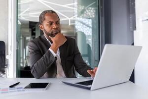 pensando maduro adulto africano americano homem de negocios às ambiente de trabalho, financista patrão investidor com computador portátil pensando sobre financeiro soluções Novo projetos dentro escritório. foto