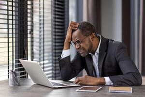 a africano homem de negocios dentro uma terno parece frustrado enquanto trabalhando em dele computador portátil dentro uma moderno escritório contexto. foto