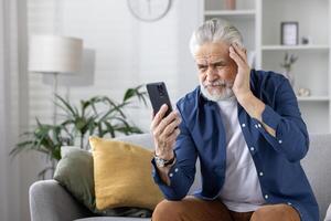 a idosos homem com uma barba parece intrigado ou preocupado enquanto tentando para usar dele Smartphone, sentado dentro uma moderno vivo sala. foto