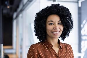 retrato do uma confiante jovem africano americano empresária sorridente calorosamente, vestido dentro uma à moda polca ponto blusa dentro uma moderno escritório contexto. foto